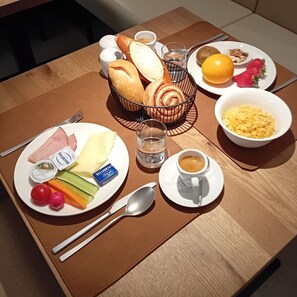 A breakfast setting with a plate of ham, cheese, and vegetables, a basket of bread, a bowl of cereal, a glass of water, a cup of coffee, and a plate with a kiwi, orange, and strawberries.