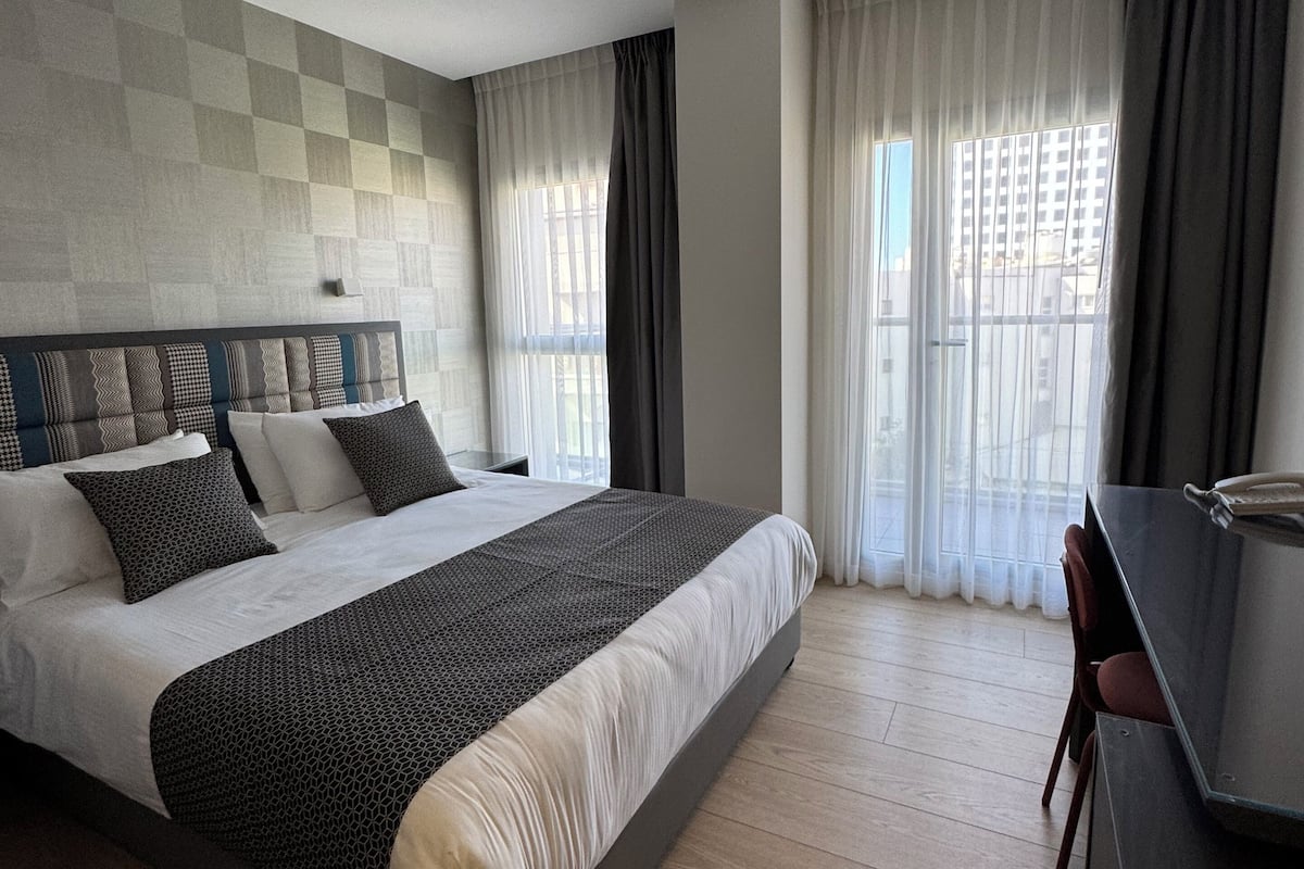 A modern hotel room with a large bed, a desk, and a view of the city through sheer curtains.