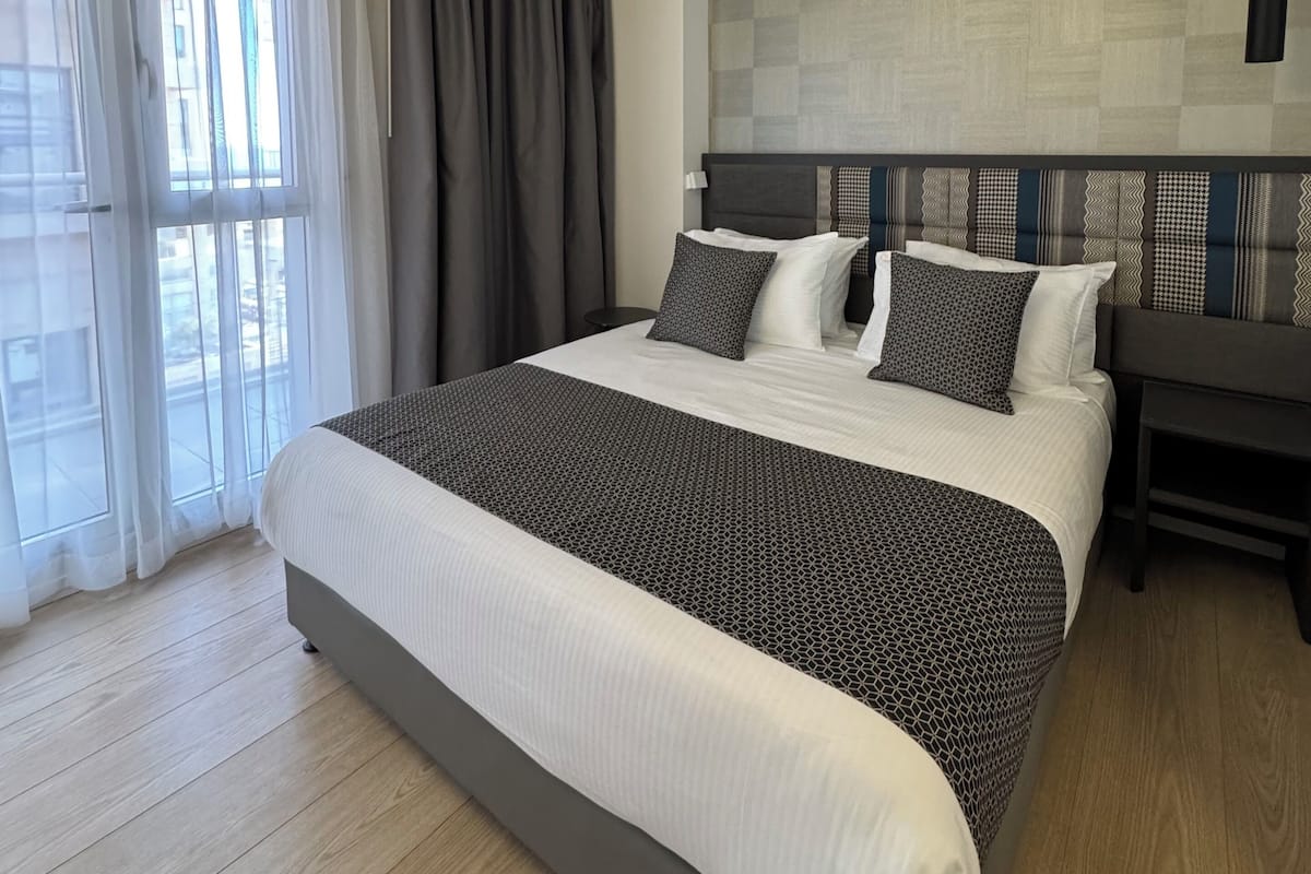 A modern hotel room with a large bed, a view of the outdoors through sheer curtains, a dark headboard, and a pendant light.