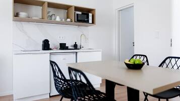 A modern kitchen with white cabinets, a microwave, and a coffee maker. There is a dining table with black chairs and a bowl of fruit.