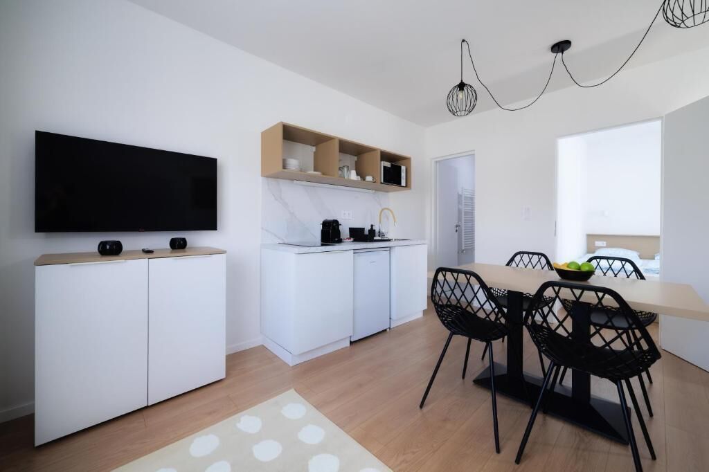 A modern kitchen with white cabinetry, a flat-screen TV, a dining area with a table and chairs, and a window providing natural light.