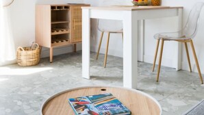Una habitación moderna y minimalista con una mesa blanca, dos sillas, un armario de madera con botellas de vino, una cesta de mimbre y una revista sobre una mesa redonda de madera.