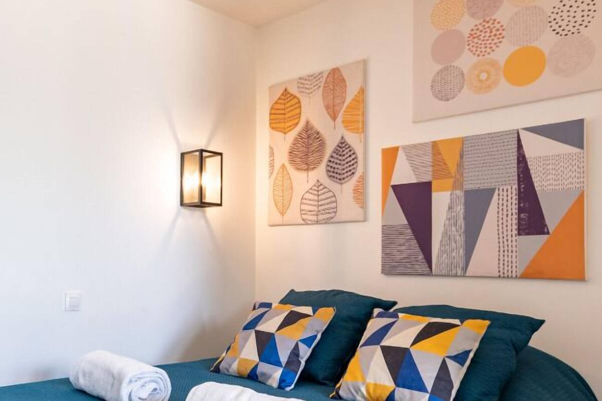 A neatly made bed with a teal bedspread and patterned pillows, a wall-mounted light, and two pieces of abstract art.