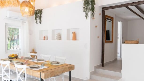 Un comedor con una mesa de madera y sillas blancas, una escalera que conduce a otra habitación, y plantas colgantes.
