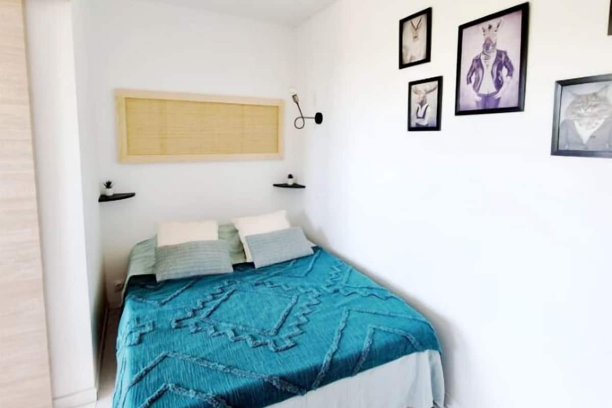 A bedroom with a bed, a window with blinds, a wall-mounted light, and several framed pictures.