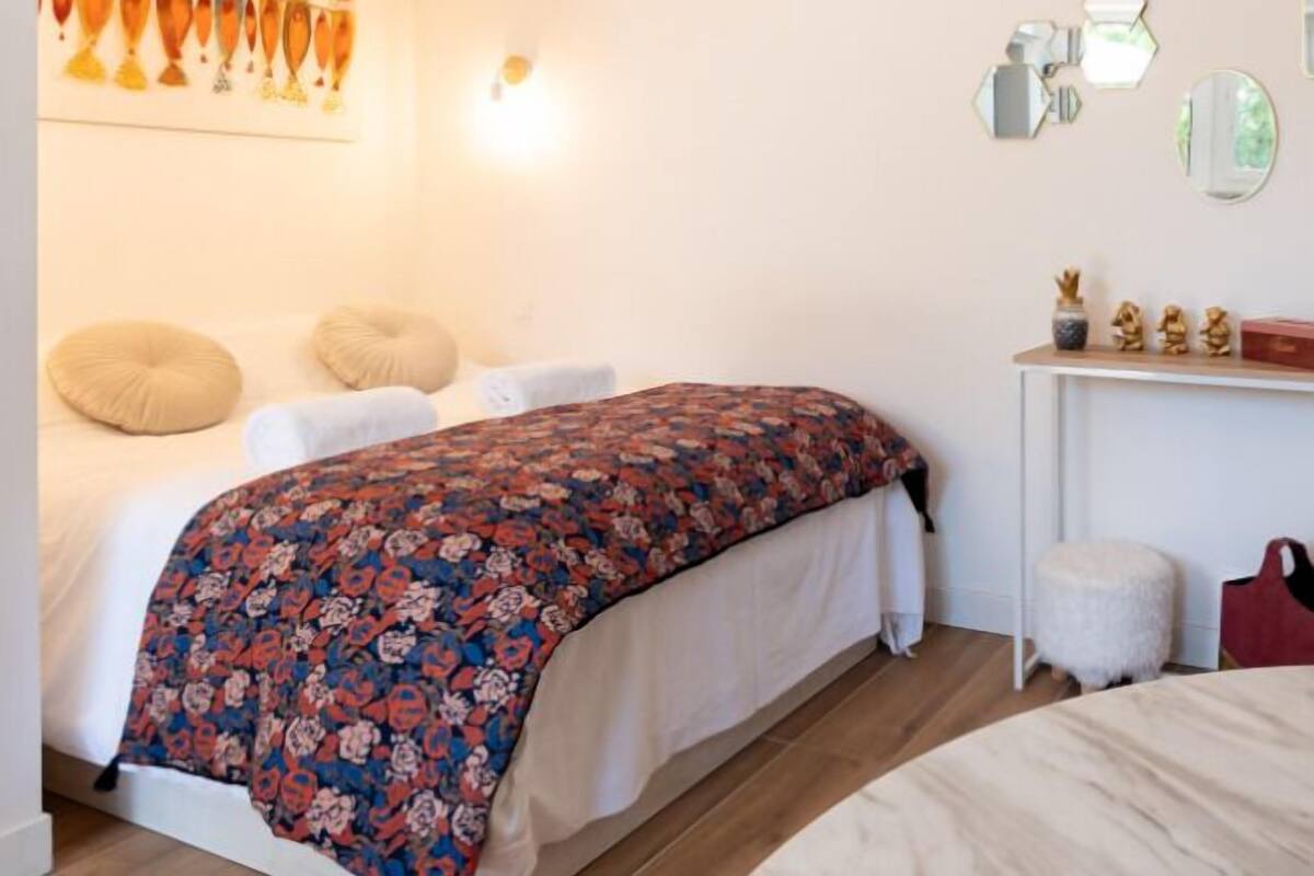 A bedroom with a bed, pillows, a bedside table with a mirror, and wall art.