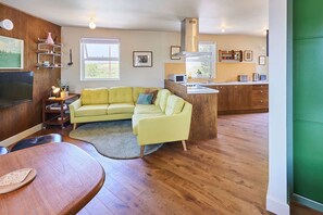 Una sala de estar con un sofá amarillo, piso de madera, una mesa de comedor con un mantel individual, un televisor y una zona de cocina con microondas y fregadero.