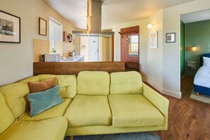 Un sofá amarillo con dos almohadas, una zona de cocina con estufa y fregadero, y un dormitorio con cama y mesita de noche.