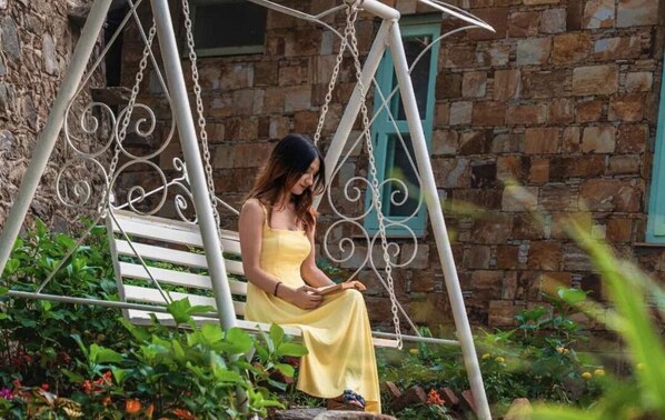 Una mujer sentada en una hamaca en un jardín con un muro de piedra y una ventana al fondo.