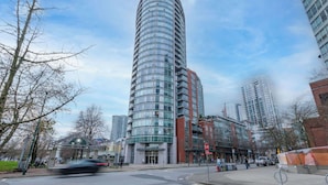 A modern high-rise building with a curved glass facade, surrounded by other tall buildings and a clear sky. - Modern Central Location with Sauna Jacuzzi & Pool (Vancouver)