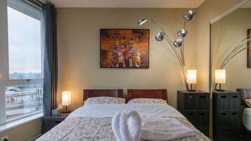 A hotel room with a bed, two bedside lamps, a painting on the wall, and a window with a view of the city.