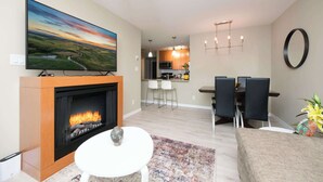 A living room with a fireplace, a flat-screen TV, a dining area with chairs and a table, and a kitchen in the background. - Modern Central Location with Sauna Jacuzzi & Pool (Vancouver)