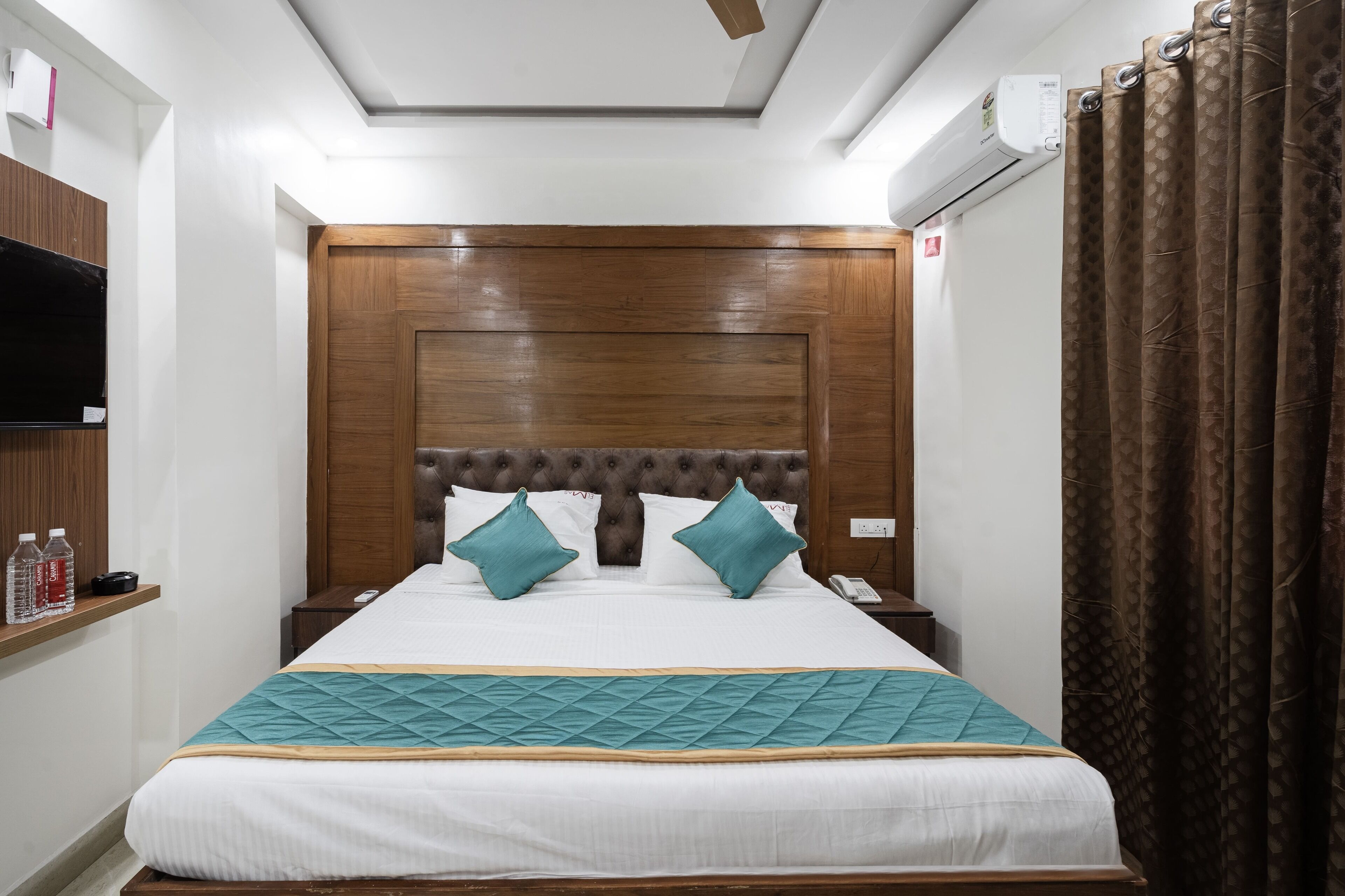 A hotel room with a wooden headboard, a bed with white bedding and teal accents, a bedside table with a phone and a bottle, and a TV on the wall.