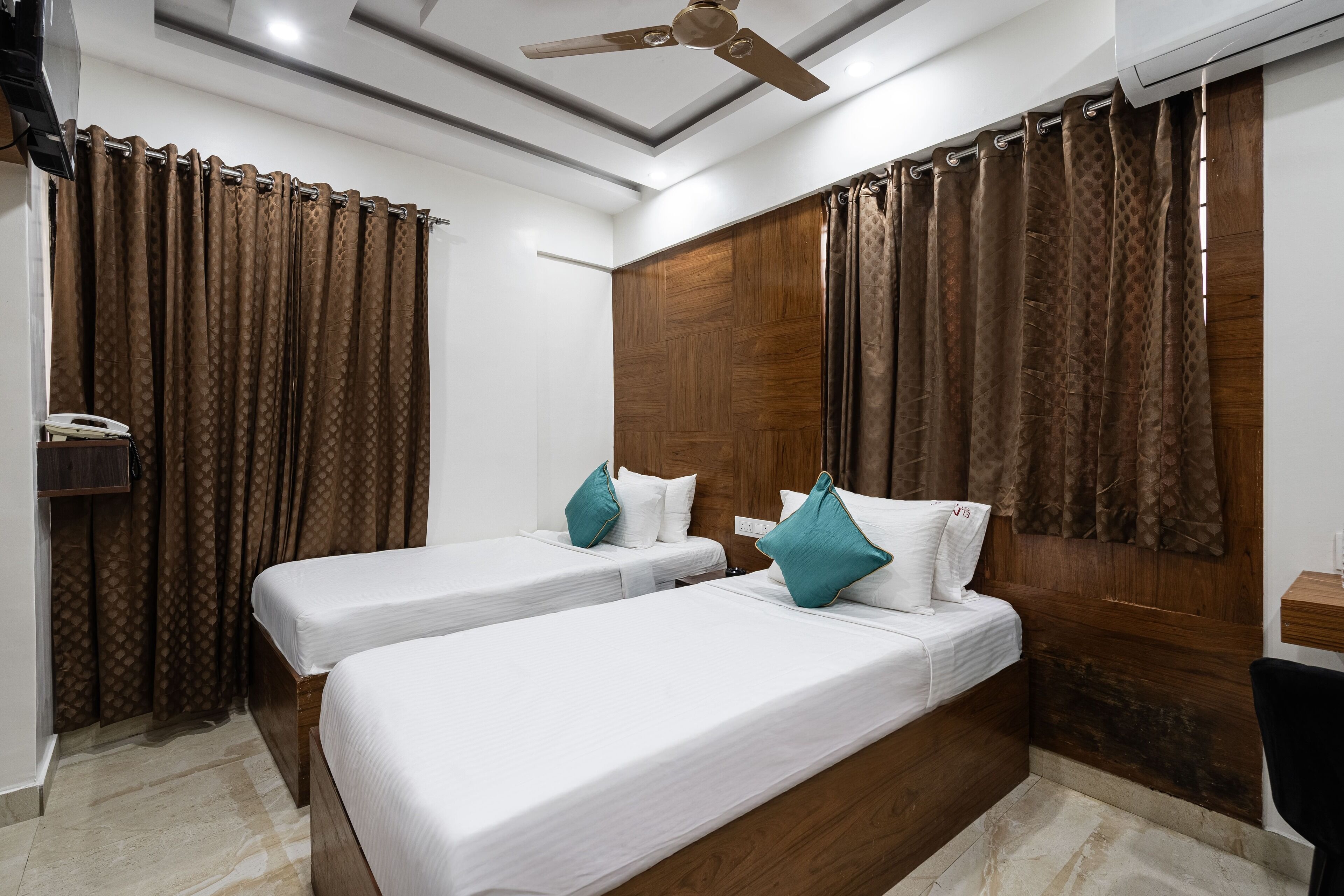 A hotel room with two single beds, wooden headboard, brown patterned curtains, a ceiling fan, and a small desk with a chair.