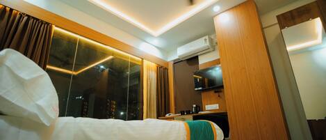 Habitación de hotel con cama, escritorio, ventilador de techo, aire acondicionado y vistas a la ciudad de noche.