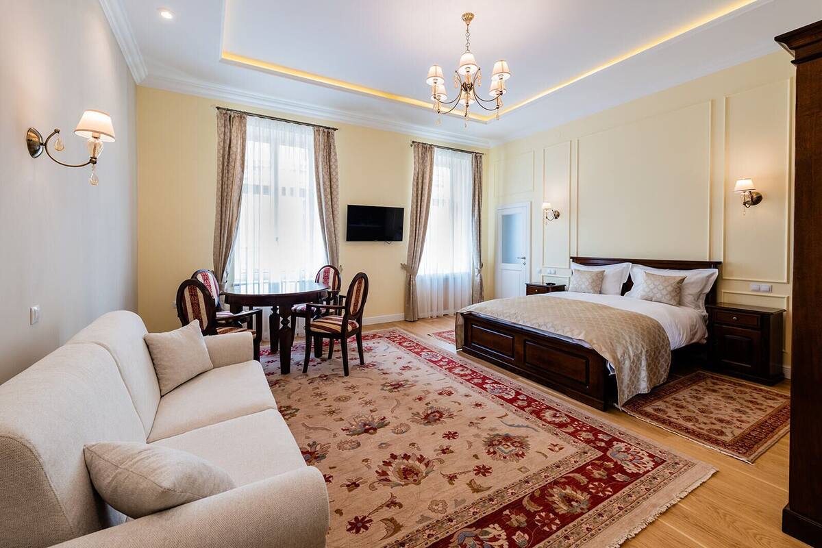 A well-lit bedroom with a large bed, a sitting area with a sofa and chairs, a dining table, and a TV mounted on the wall.