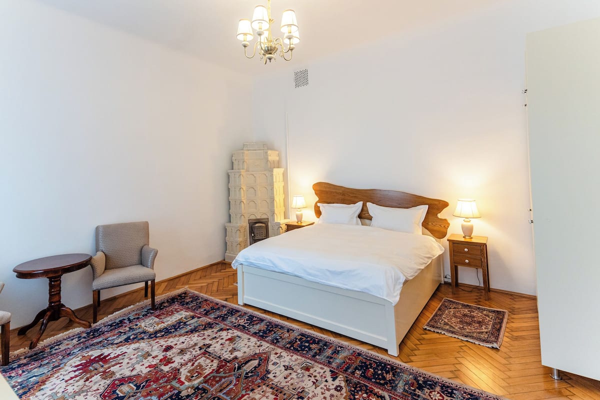 A bedroom with a bed, a chair, a nightstand, a lamp, a chandelier, and a patterned rug.