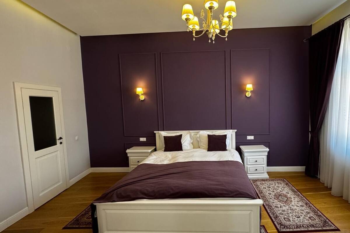 A bedroom with a large bed, two bedside tables, a chandelier, and a window with curtains.