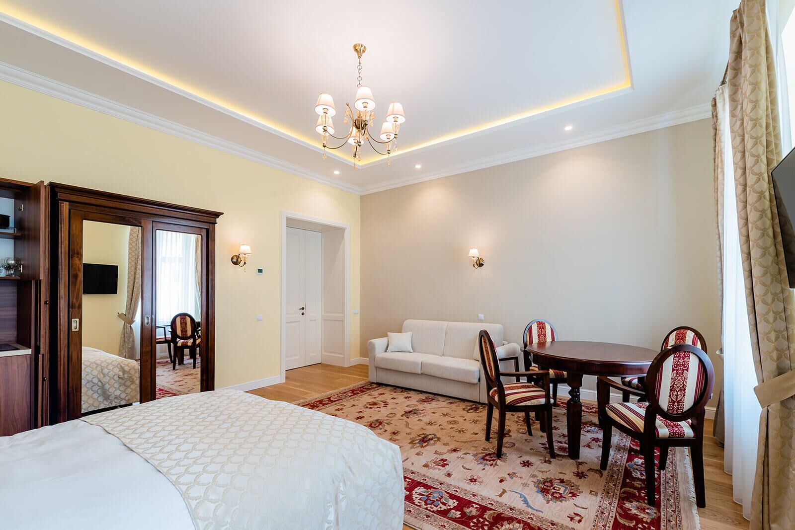 Ein Hotelzimmer mit einem Bett, einem Sofa, einem Esstisch mit Stühlen, einer Chandelier und einem Spiegel.