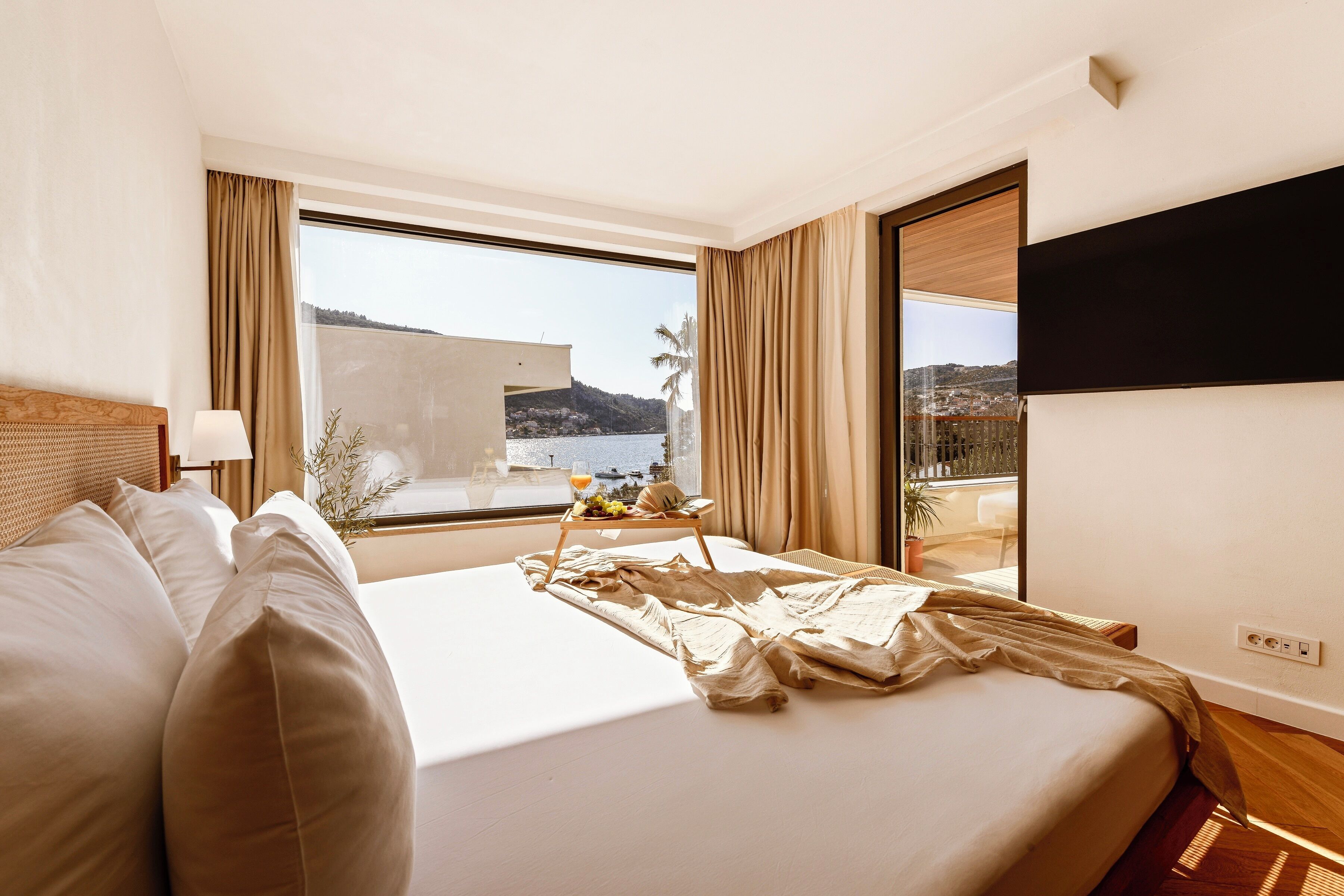 A hotel room with a large bed, a TV mounted on the wall, and a window offering a view of the sea and hills.