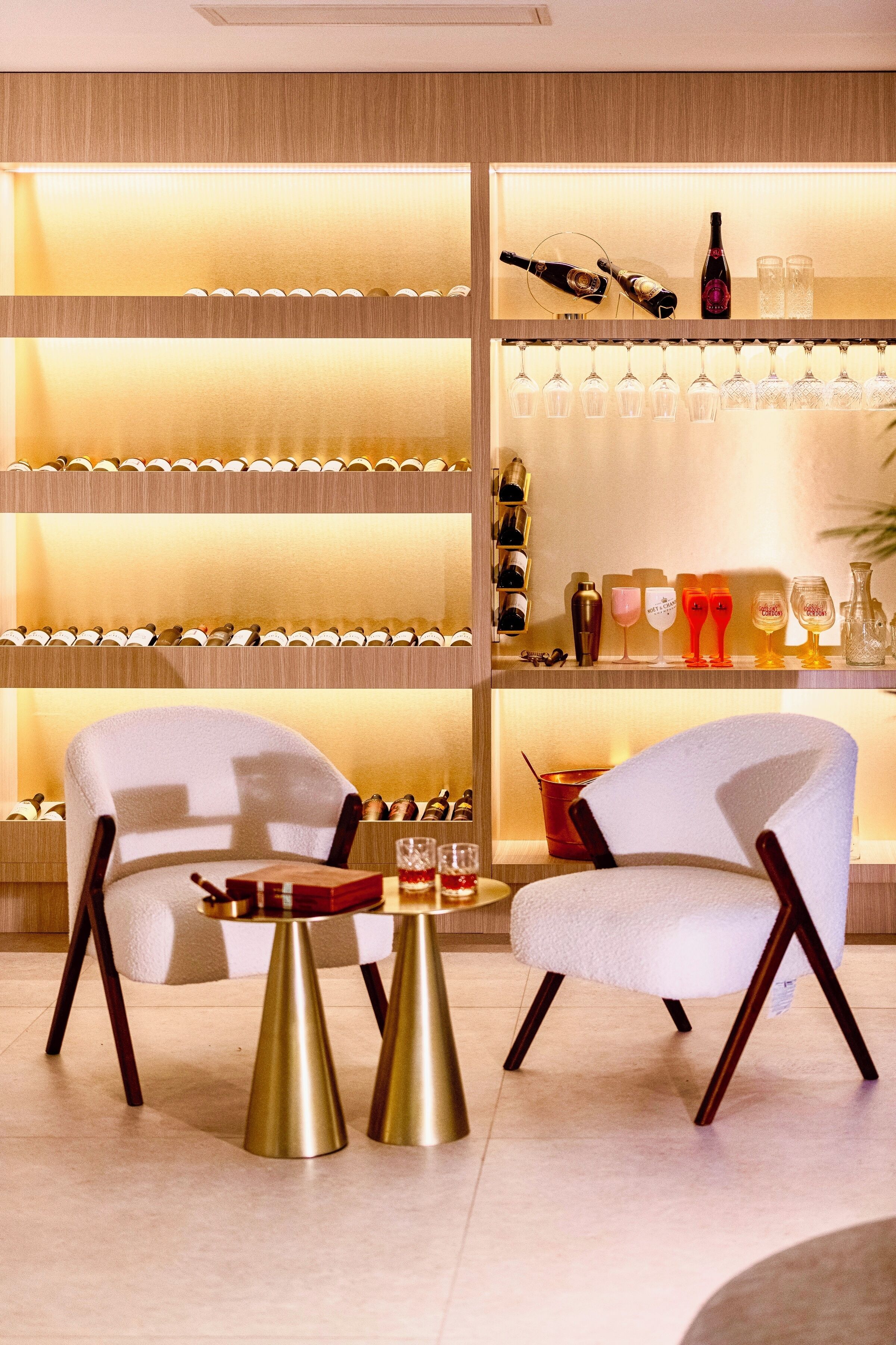 Un intérieur moderne avec deux fauteuils blancs, une table dorée et des étagères remplies de bouteilles de vin et de verres.