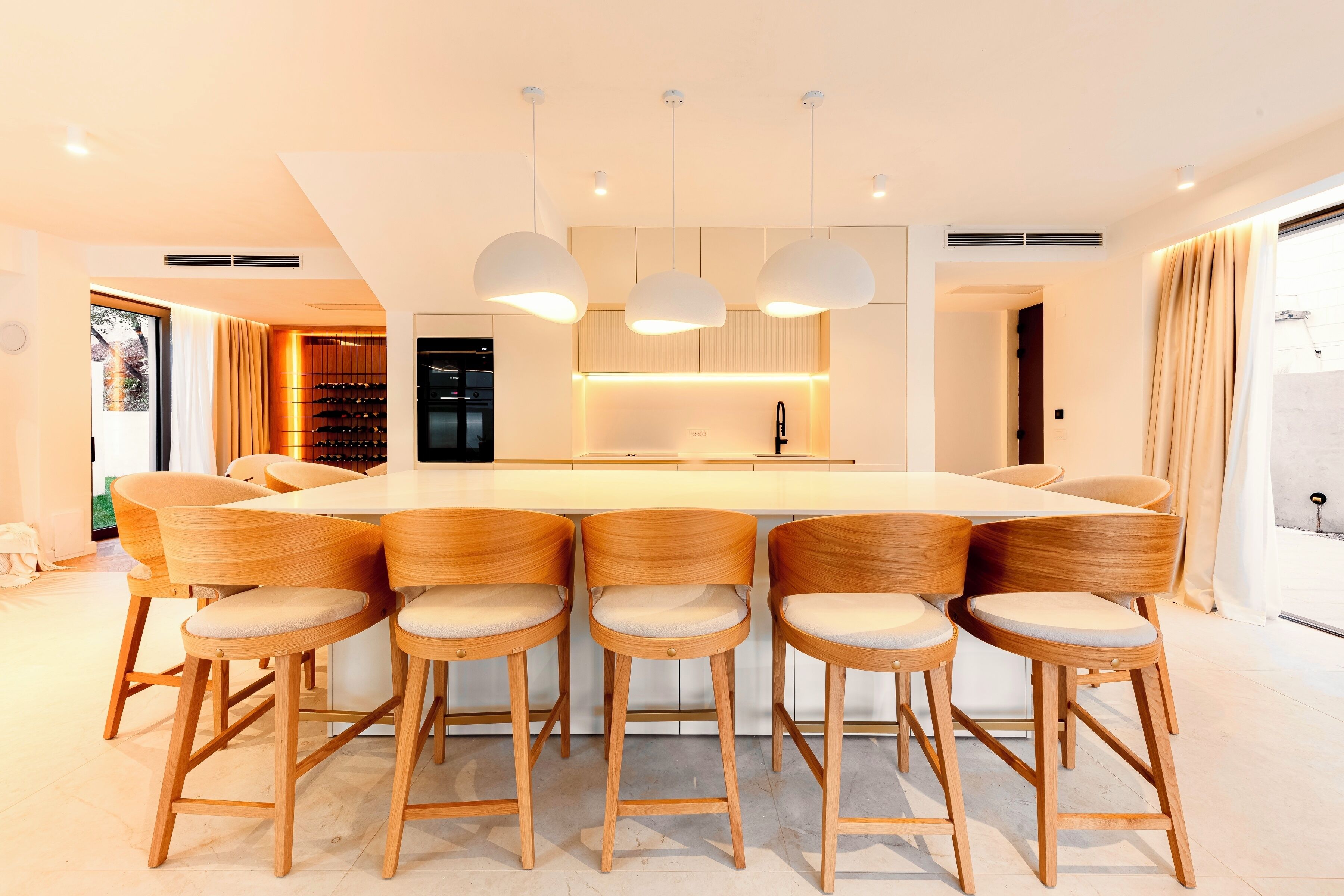 A modern kitchen with a central island, wooden bar stools, pendant lighting, and a view of the outdoors.
