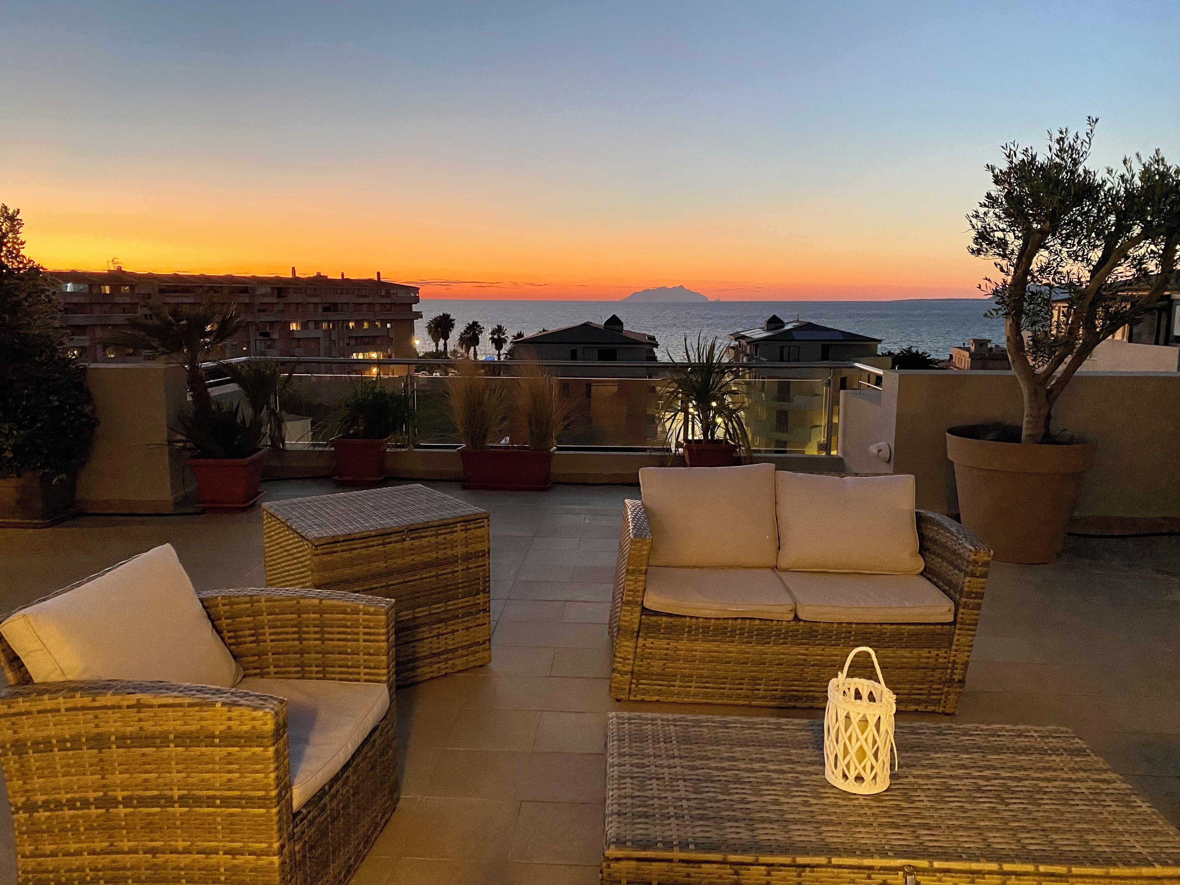 A terrace with wicker furniture and a lantern, overlooking a sunset and a body of water.