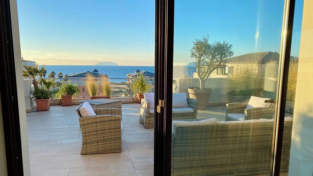 Ein Balkon mit Korbmöbeln und Blumentöpfen, der Blick auf eine Küstenlandschaft mit Gebäuden und Bergen freigibt.
