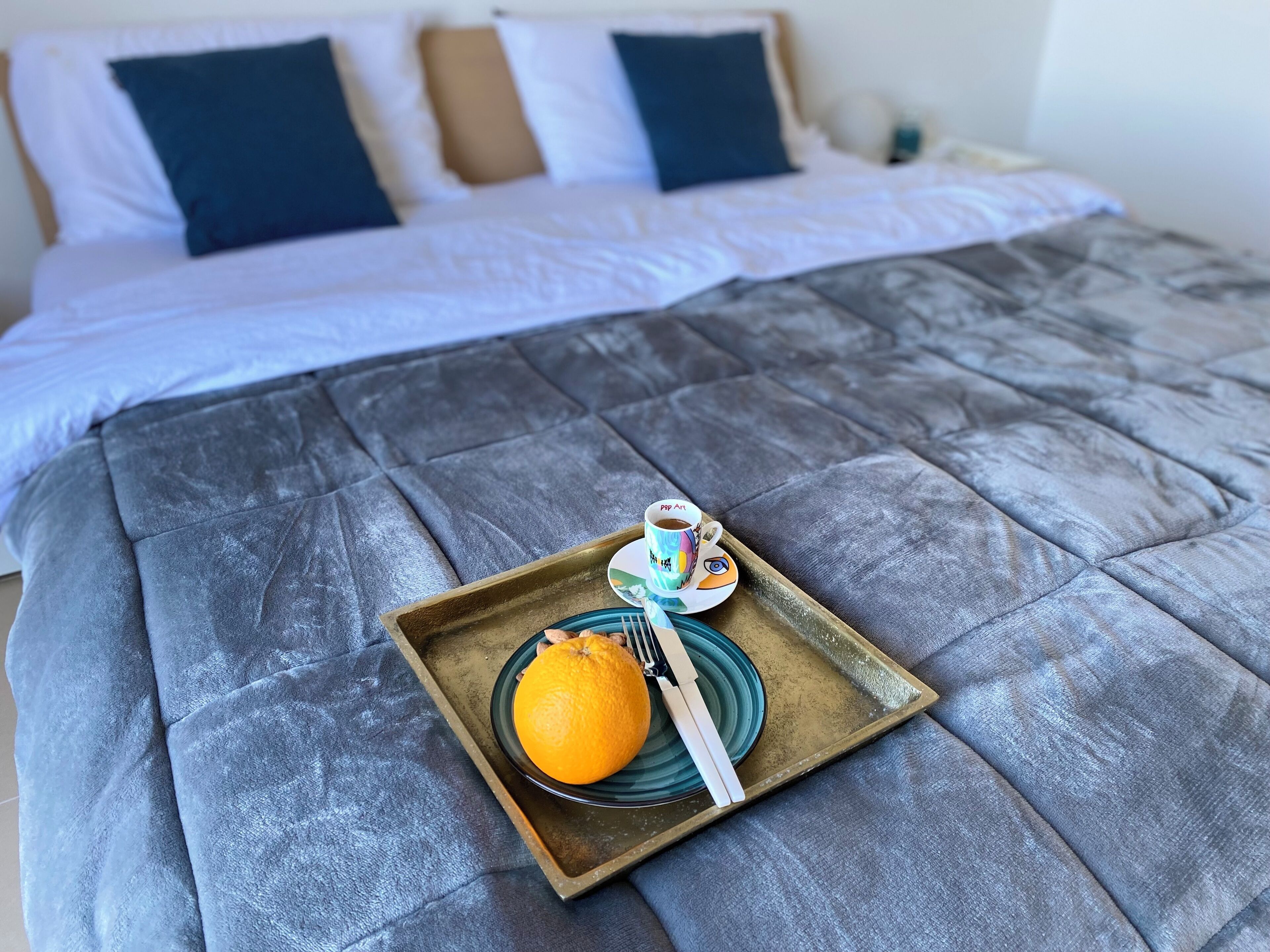 Un letto con una coperta grigia, un vassoio con un'arancia, una tazza e un cucchiaino.