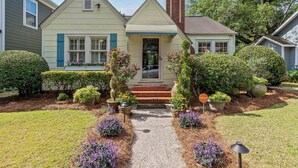 A two-story house with a front porch, a brick chimney, and a well-maintained garden with various plants and flowers. - La Petite Fleur Rouge | Elegant & Cozy 2/1 Getaway (North Charleston)
