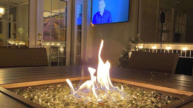 A fire feature with glass tiles, a television mounted on the wall, and a view of a building with large windows. - La Petite Fleur Rouge | Elegant & Cozy 2/1 Getaway (North Charleston)