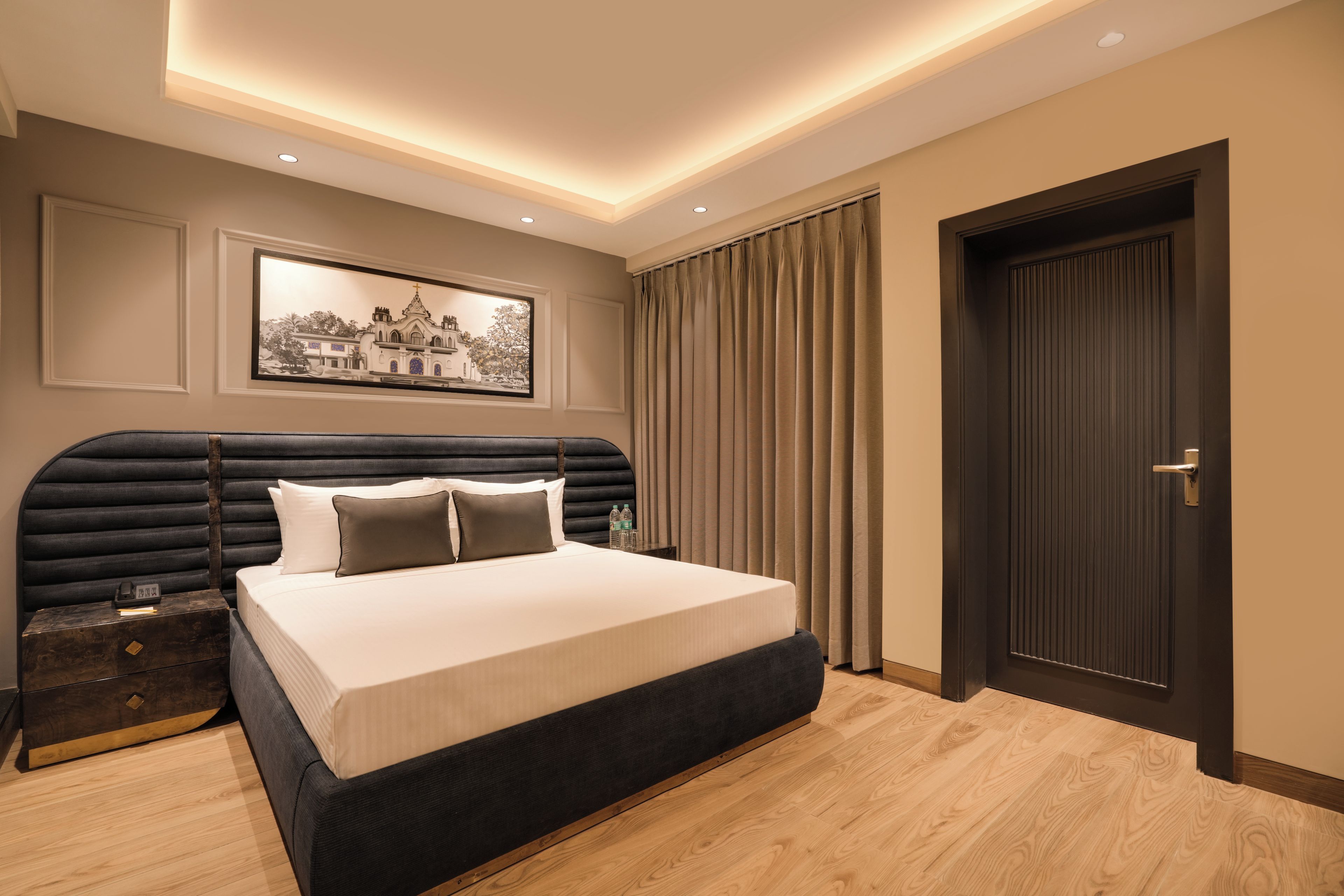 Quarto de hotel moderno com cama grande, mesa de cabeceira, quadro na parede e uma porta.