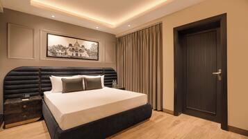 Quarto de hotel moderno com cama grande, mesa de cabeceira, quadro na parede e uma porta.