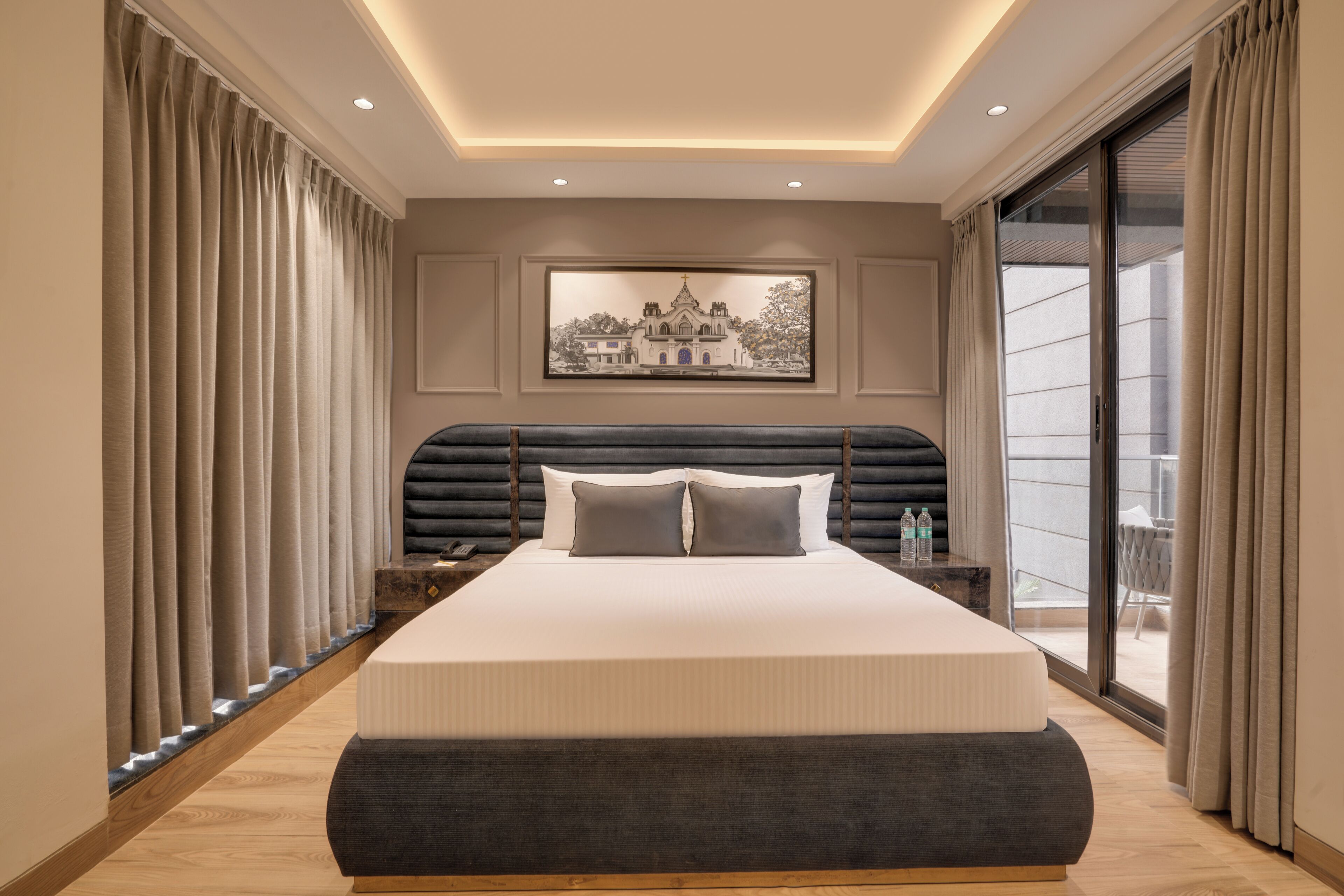 A modern hotel room with a large bed, a view of a building through the window, and a balcony with curtains.