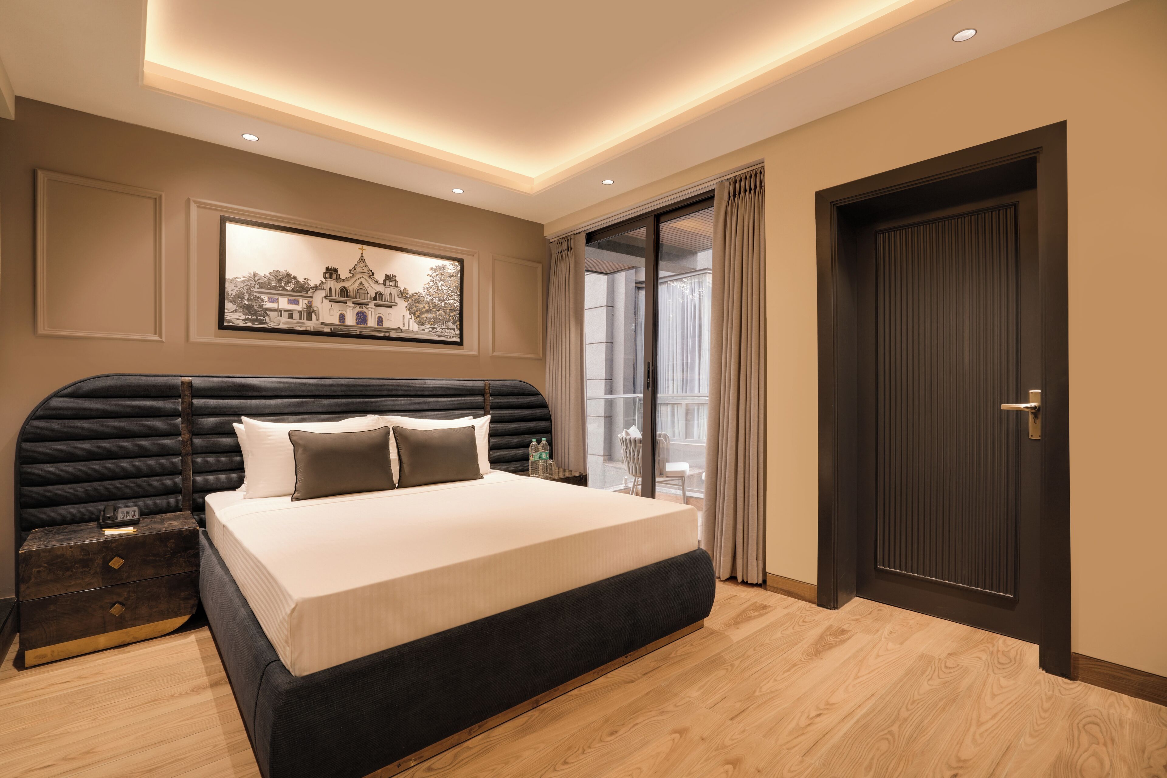 Ein modernes Hotelzimmer mit einem großen Bett, einem dunklen Kopfteil, einem Nachttisch mit einer Uhr, einem gerahmten Bild an der Wand und einer Tür rechts.