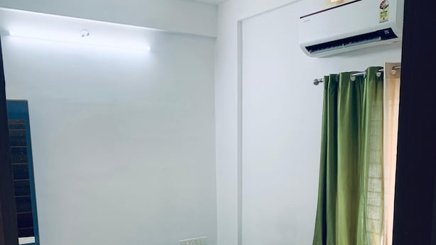 A neatly made bed with a pillow and a brown item on it, a ceiling fan, an air conditioning unit, and a green curtain partially drawn. - Celestia residency (Kanayannur)