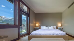 Ein Schlafzimmer mit einem großen Bett, zwei Nachttischen mit Lampen, Blick auf Berge durch ein Fenster und einem Teppichboden.