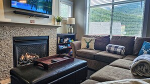 A living room with a fireplace, a television mounted on the wall, a sofa, and a window with blinds. - Ski-In/Ski-Out Mountain Retreat / Hot Tub + Garage (Panorama)