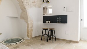 A small kitchen with a white wall, a black countertop, two stools, and a window with a curtain.
