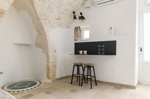 A small kitchen with a white wall, a black countertop, two stools, and a window with a curtain.
