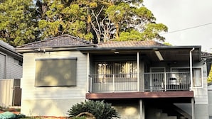 A single-story house with a balcony, a stone wall in front, and a large tree in the background.