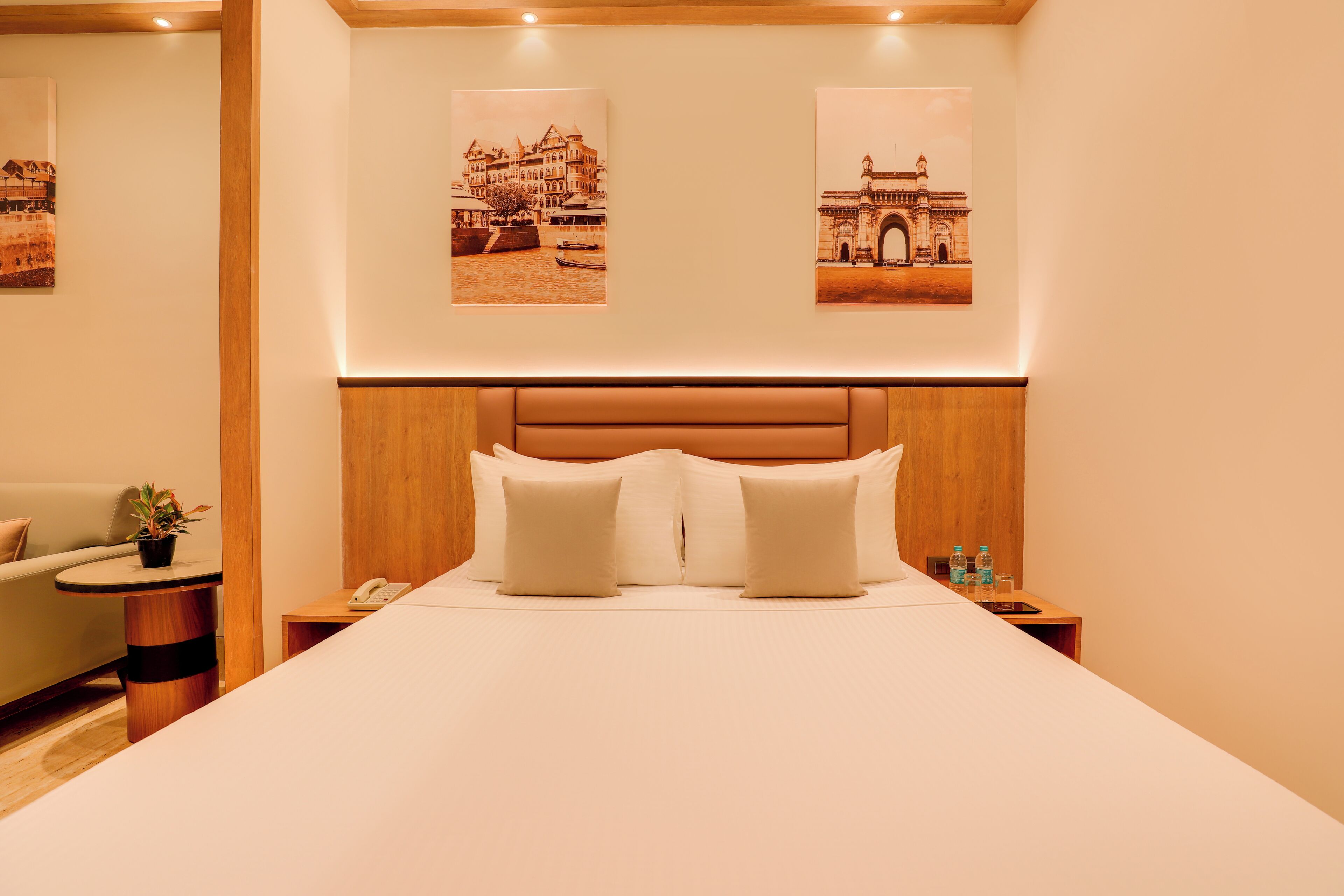 A hotel room with a large bed, wooden headboard, two bedside tables with a phone and water bottles, a small side table with a plant, and two framed pictures on the wall.