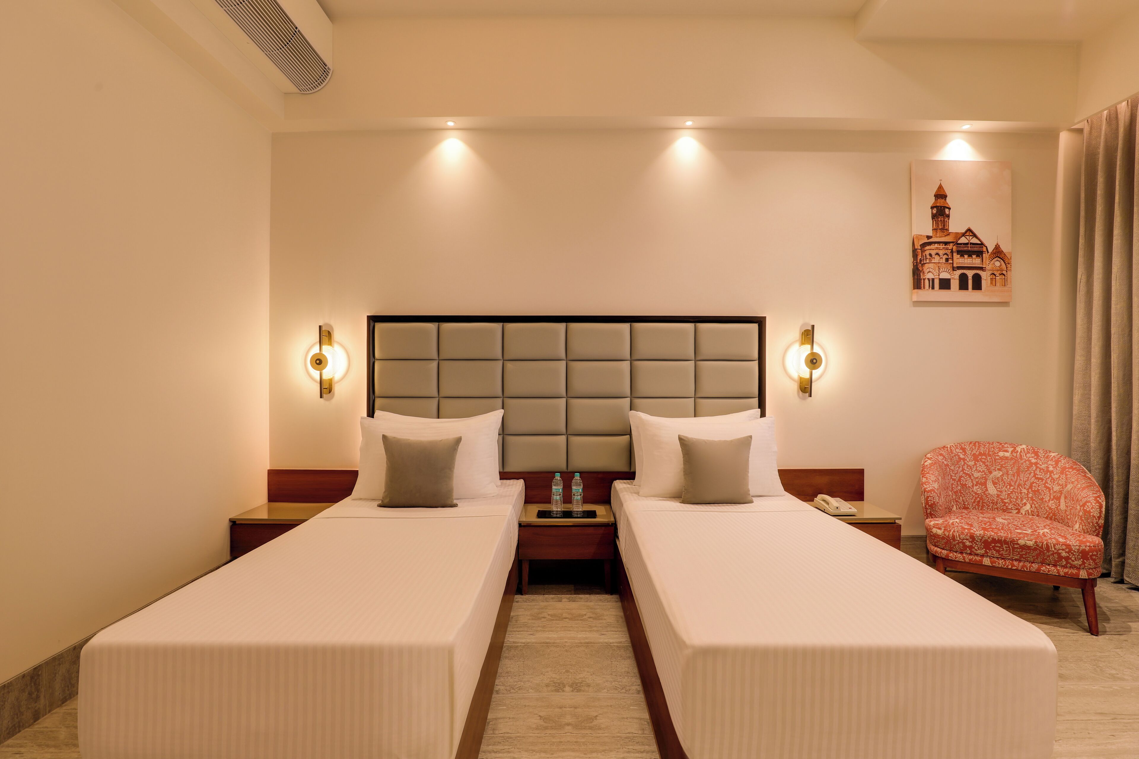 A hotel room with two single beds, a headboard, bedside tables, a chair, and a wall-mounted artwork.