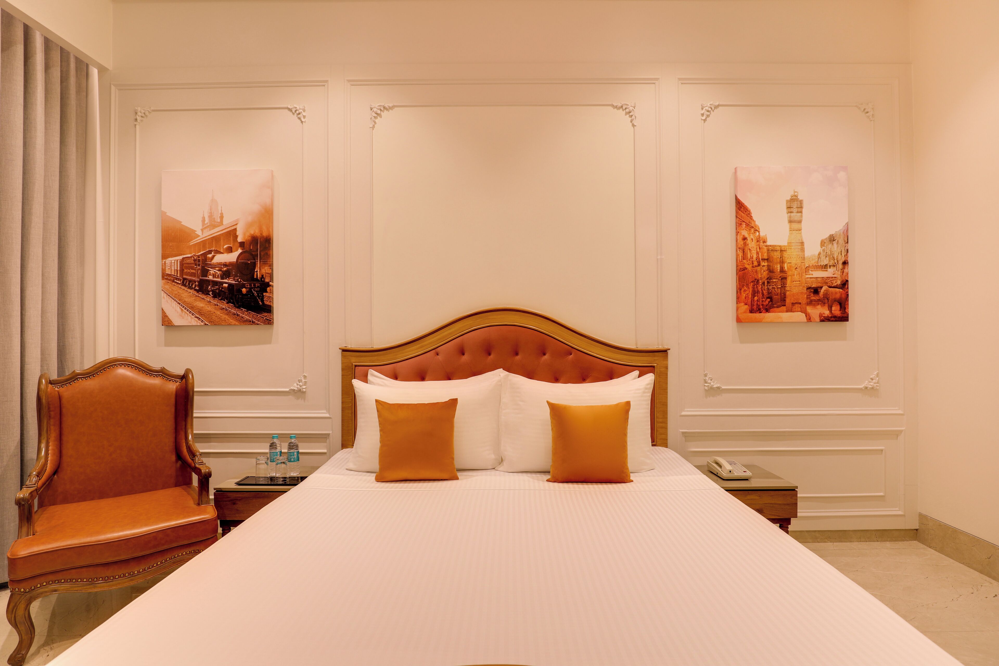 Une chambre d’hôtel avec un grand lit, une chaise en bois, une table de chevet avec une lampe, et deux tableaux encadrés au mur.