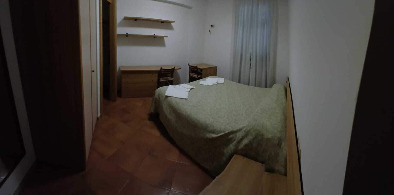 A small, dimly lit room with a single bed, a wooden dresser, and a desk with a chair.