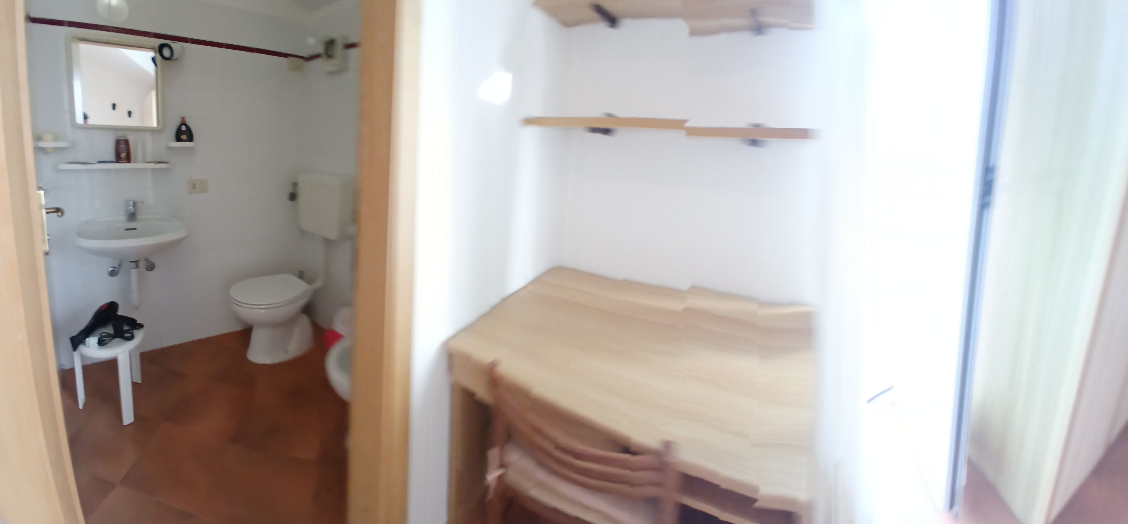 Ein Badezimmer mit weißem Waschbecken, Toilette und Spiegel. Ein kleiner Hocker mit Gegenständen darauf. Der Boden ist mit Fliesen ausgelegt.