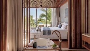 A modern bedroom with a large bed, a bedside table with a bottle and glass, and a view of a beach and palm trees.