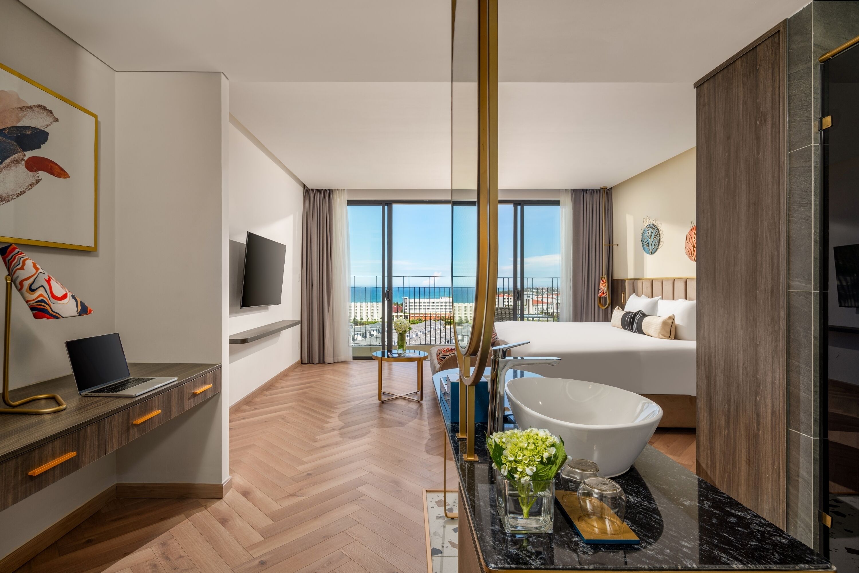 Habitación de hotel moderna con una cama grande, una zona de estar con una computadora portátil, un ventanal con vista a la ciudad y un baño con una bañera espaciosa.