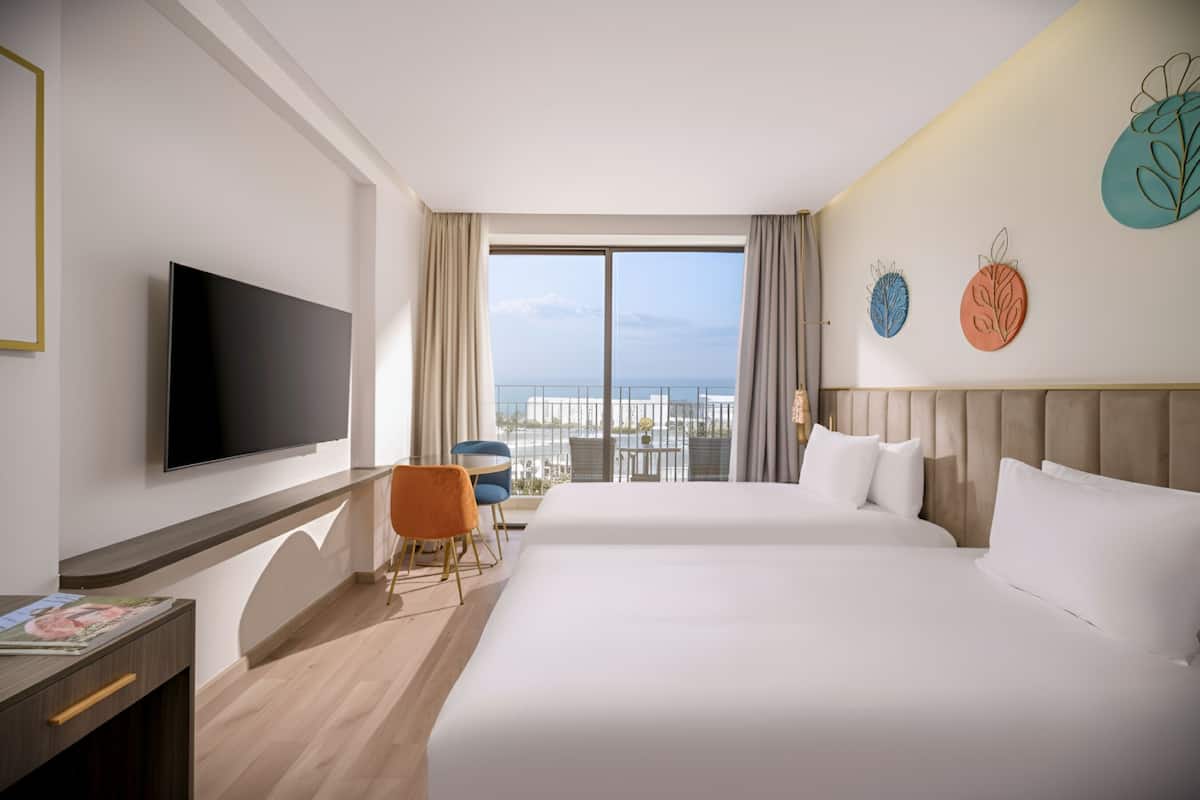 A hotel room with a large bed, a TV, a desk, and a balcony with a view of the sea.
