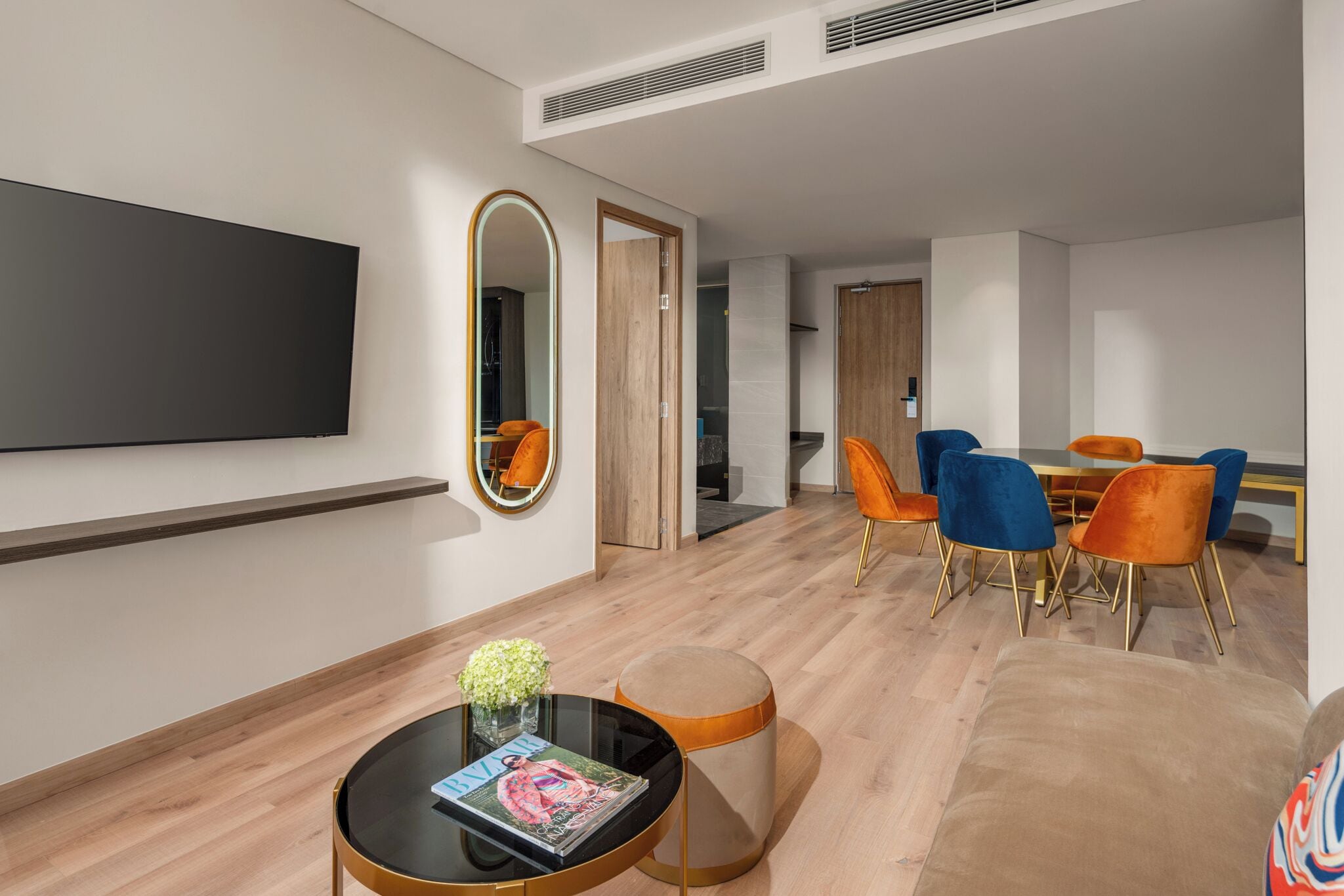 A modern living room with a flat-screen TV, a round coffee table with a magazine, and a dining area with orange and blue chairs.